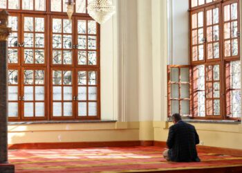 Pria duduk berdoa di interior masjid dengan cahaya alami, ilustrasi qunut subuh
