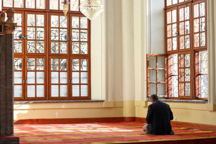 Pria duduk berdoa di interior masjid dengan cahaya alami, ilustrasi qunut subuh