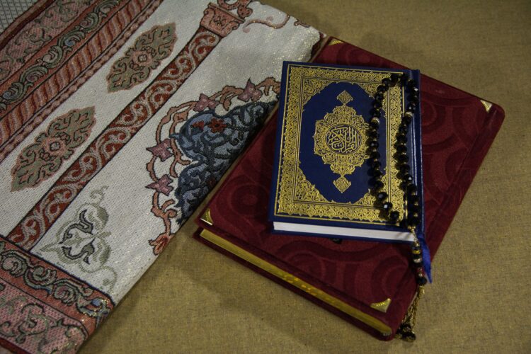 Mushaf Al-Qur'an bersampul biru emas dengan tasbih hitam di atas sajadah, ilustrasi keutamaan dzikir tasbih subhanallah