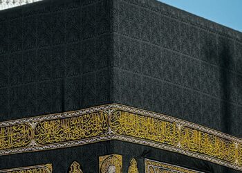 Close-up sudut Ka'bah dengan kaligrafi emas dan menara masjid di latar langit biru, ilustrasi keutamaan mencium Hajar Aswad