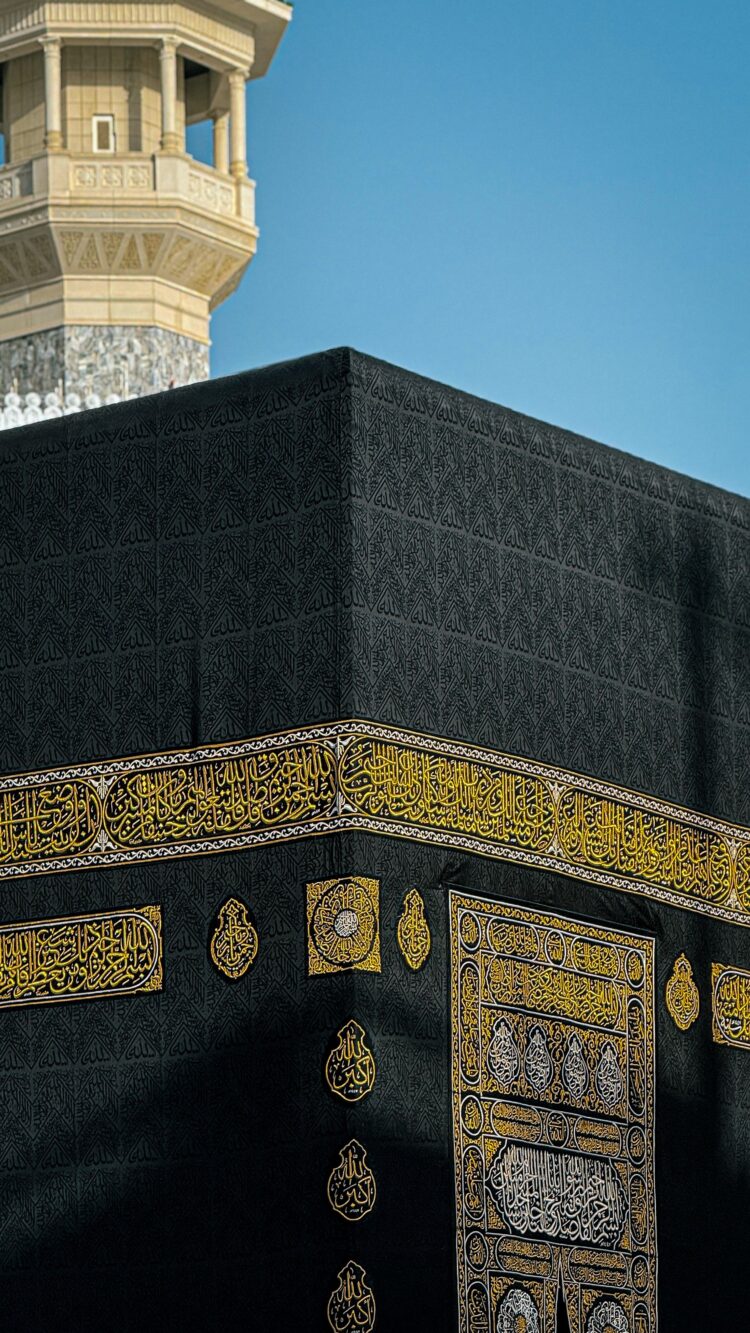 Close-up sudut Ka'bah dengan kaligrafi emas dan menara masjid di latar langit biru, ilustrasi keutamaan mencium Hajar Aswad